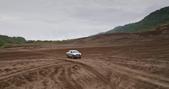 Pickup truck driving on a vast, rugged volcanic ash plain with a green hillside in the background under an overcast sky, showcasing off road adventure and exploration in Vanuatu. Drone flight