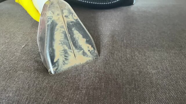 A close-up of a washing vacuum cleaner attachment on brown upholstered furniture. The nozzle shows dirt and debris being extracted from the fabric surface.