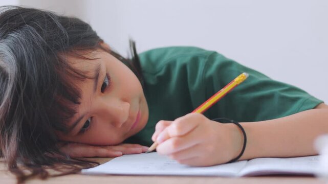 Close up selective focus of a tired Asian kid girl resting her head on the desk. Stressed and exhausted from studying too much homework in bright daylight. Education burnout and overwork concept.