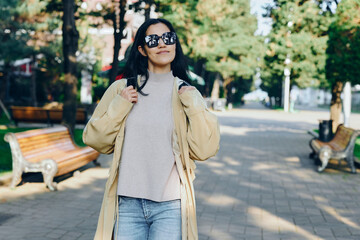 Obraz premium Woman in a beige coat and gray sweater walks in a sunlit park, showcasing casual street fashion and outdoorsy confidence in an urban setting