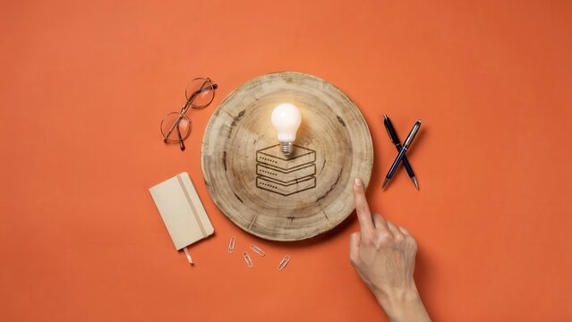A hand holds a plate with cake and light bulb next to glasses and notebook on table