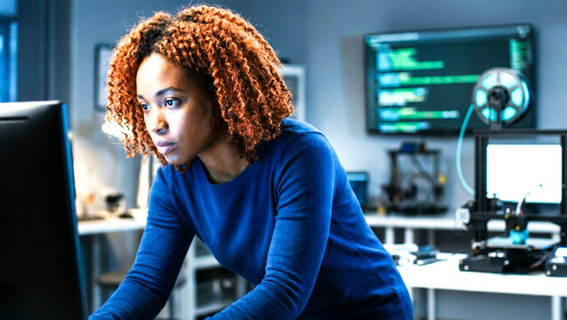 A woman with curly hair wearing a blue shirt working on a computer in a modern office with coding and 3d printing equipment