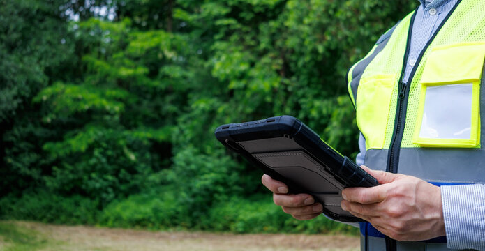 Forestry engineer or ecologist holding digital tablet in protective rugged case during field research in green forest. Data logging, environmental monitoring and tree mapping outdoor