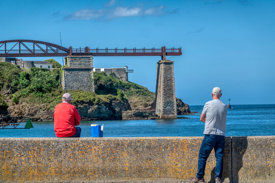 Two men relaxing on a concrete pier in Ribadeo, one fishing in a red sweater, with the historic iron structure of O Cargadeiro over the blue sea in the background
