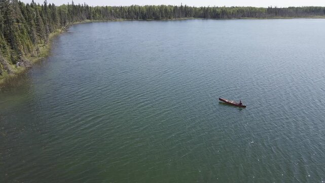 Drone flying over a person in a red canoe while floating on a small northern lake that is surrounded by an evergreen forest

