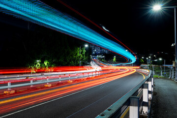 Long exposure shot capturing colorful light trails from vehicles on a curved road at night, with glowing cyan LED structure overhead and city lights in the background, South Korea. © CHO KEE HO