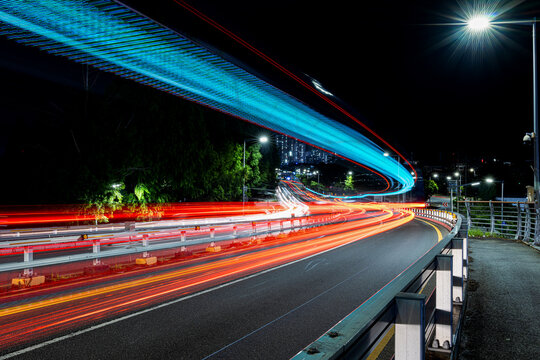 Long exposure shot capturing colorful light trails from vehicles on a curved road at night, with glowing cyan LED structure overhead and city lights in the background, South Korea.