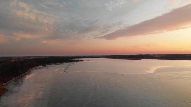 POV from a drone flying above a small lake covered in ice as the evening creates an orange sky
