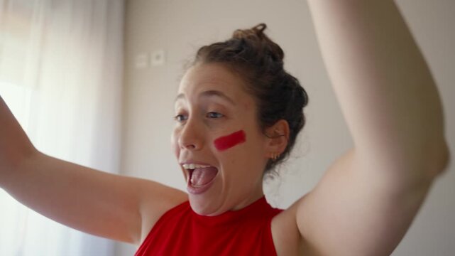 Enthusiastic female sports fan is at home watching a game celebrates in joy. Young woman in red colors and face paint excited cheerful during game time. Soccer football or basketball fan in apartment