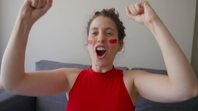 Female sports fan dressed in red watching game on TV celebrating after a goal. Young woman enjoys soccer or football match shouts goal with hands in the air in joy cheering in victory in living room