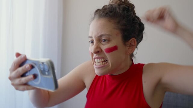 Passionate sports fan watches live game streaming on her smartphone celebrating. Woman with red face paint and clothes of favorite team excited by result or play in the match. Shouting cheering in joy