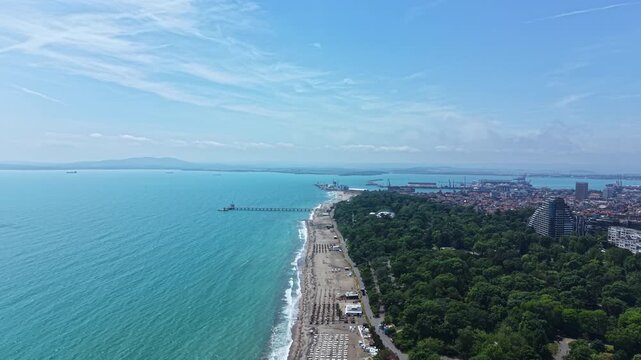 Clear blue water meets a sandy beach with green trees along the shore. People enjoy the day by the water, while ships sail in the distance. The setting shows a lively beach scene.