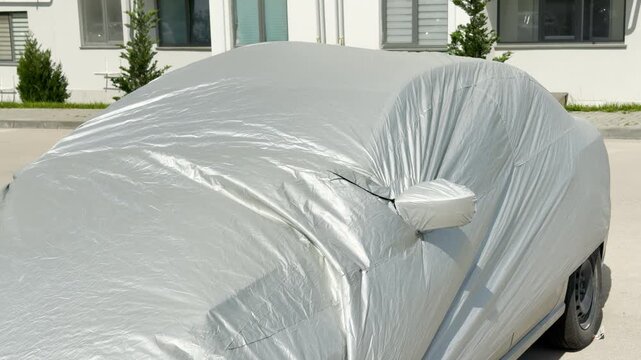 A silver waterproof cover completely blankets a parked car. The automobile stands protected under bright sunlight in an asphalt parking lot next to a white building.