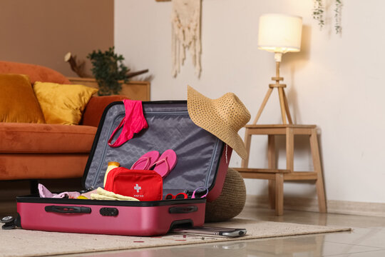 Suitcase with first aid kit, stylish clothes and summer hat on floor in room