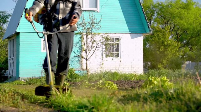 Man mows lawn with grass trimmer near house. Bright blue house stands behind him in background. Green grass and trees surround the yard. He works methodically, focused on his task