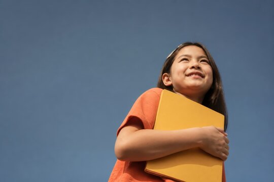 Smiling young girl holding a yellow folder outdoors against a clear blue sky in a carefree lifestyle moment