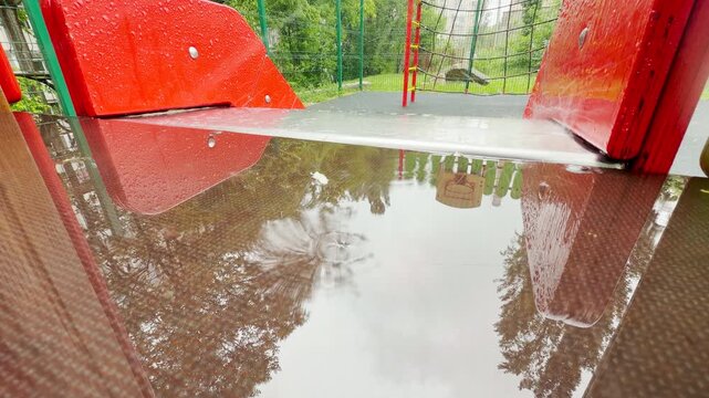 rain hits a playground surface, creating a quiet splashing sound. The wet red structure feels lonely yet peaceful under the grey sky.