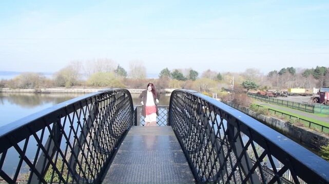 Slow motion close shot of a woman walking across a railway bridge on a bright spring day with trees and a river in the background