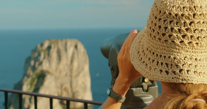Close view of woman with straw hat using telescope toward Capri sea stacks, Italy