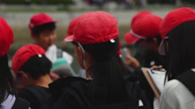Group of school children students outdoor excursion, guided tour in the city with guide, docent with tourist pupils visitors, wear color caps hats, kids class field trip, urban sightseeing in Japan