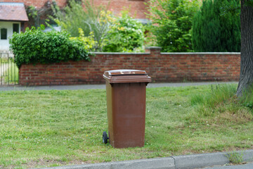 Naklejka premium A garden container for bio-waste and garden debris stands on the lawn. It features two rubber wheels for mobility. A blurred brick wall and greenery are in the background.