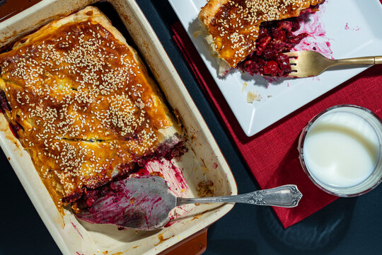 Homemade berry pie with sesame seeds and a glass of milk, closeup. Top view of sweet cherry pastry pie in a baking dish next to a slice on a plate and a glass of milk