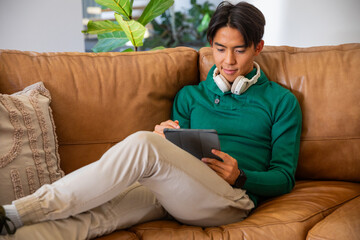 Obraz premium Asian man sitting on tan leather couch in living room holding dark tablet wearing headphones
