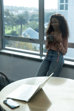 African-American woman leaning on sill in pink blouse jeans holding gray mug by laptop, copy space