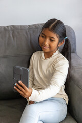 Obraz premium Indian school-age girl sitting on grey sofa at home holding grey-cased smartphone and smiling