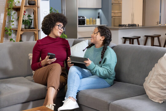 African-American mother in burgundy top and daughter sitting on sofa at home with phone and tablet