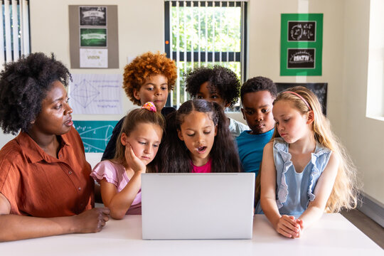 Diverse students and African American teacher in rust shirt gathering around laptop in classroom