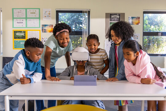Diverse children gathering around seated student wearing white VR headset, watching tablet in class