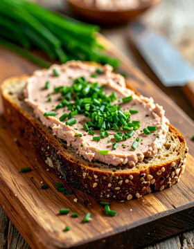 Fresh slice of multigrain bread with savory meat pate and chopped chives, metal knife on rustic wooden board, appetizing gourmet snack, tasty food. Close-up.