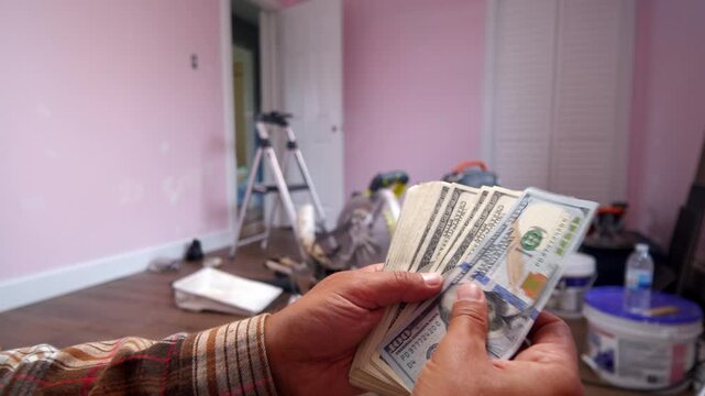 A man carefully counts dollar bills, planning for home renovation materials. The room is cluttered with tools and paint, showcasing a project in progress