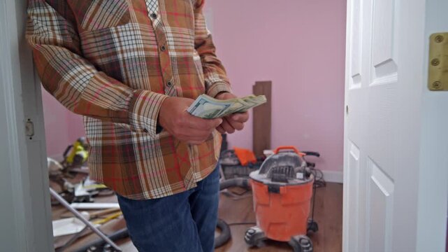 A man is seen carefully counting dollar bills to determine how much money to allocate for materials needed to renovate his home, surrounded by tools