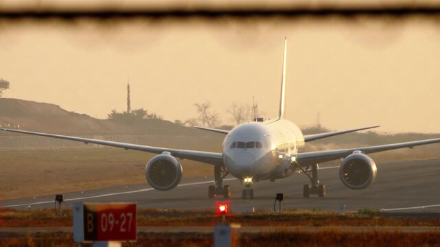 Commercial passenger airplane taxiing on runway during golden hour sunrise with engine heat haze, front view telephoto lens shot