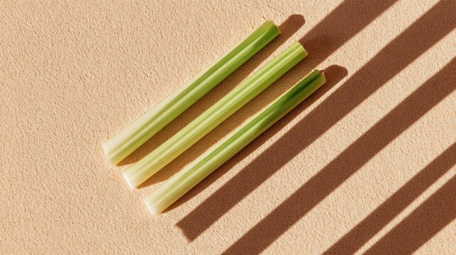 A minimalist composition of three celery sticks placed parallel on a pastel sand background with long directional shadows