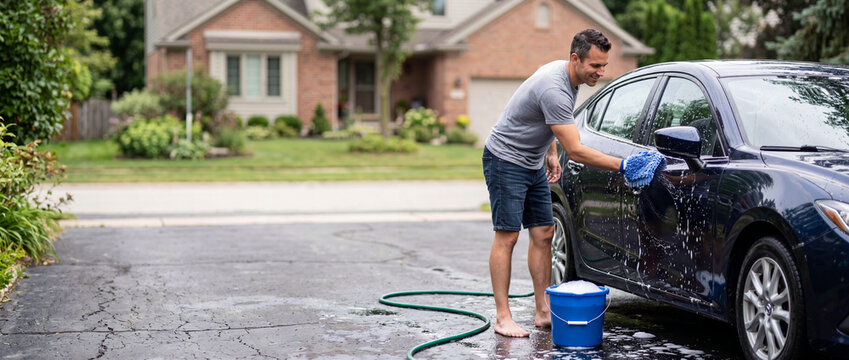 Smiling man washing his dark blue car in the driveway. Barefoot adult male cleaning vehicle with soapy mitt and bucket outside a suburban house. Home maintenance chore
