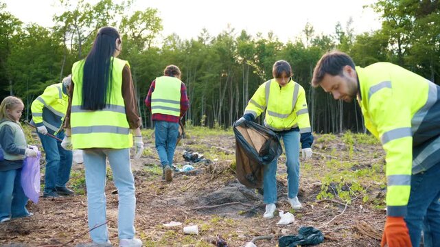 Adults and child collecting litter on cleared land. Eco volunteers wearing safety vests and holding waste bags. People cleaning nature area together. Community environmental cleanup activity.
