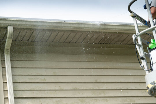 Man in the bucket of a lift, cleaning vinyl siding on second story of a house