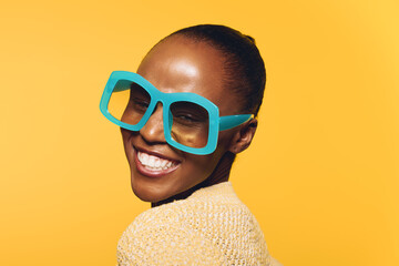 Happy dark skinned African woman smiles wearing large blue sunglasses and beige sweater, posing on...