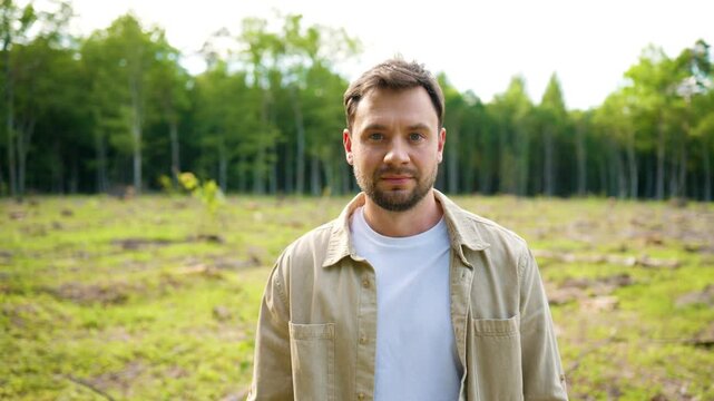 Caucasian man standing on deforested land after tree cutting. Male looking at camera near cleared woodland area. Environmental impact and forest loss concept. Logged forest site.