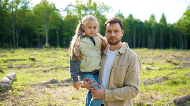 Caucasian father holding little daughter on deforested land and looking at camera. Family witnessing consequences of logging and habitat loss. Environmental protection and sustainable future concept.