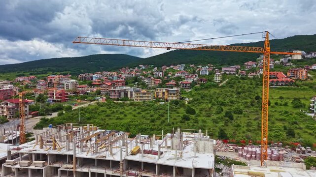 A construction site shows a large crane over a building under development. Nearby, several houses and green hills are visible. The weather is cloudy with no rain.