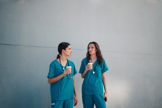 Female medical staff nurses discussing during coffee break, wearing scrubs outside hospital building