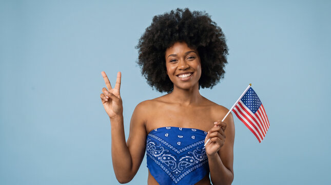 Black young woman with afro hairstyle holding American flag and showing peace sign on soft blue background, patriotic 4th of July portrait celebrating Independence Day in USA
