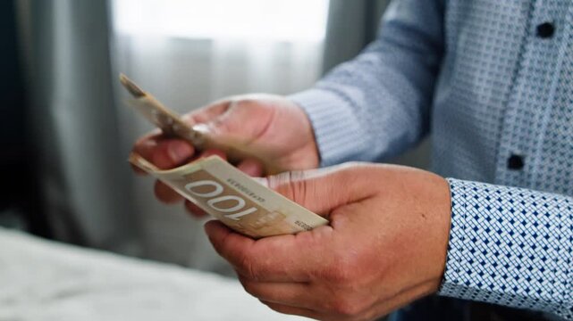 A man focuses on counting Canadian dollars with diligent hands in a cozy room. Soft light highlights the intimate moment, revealing the value of each note and time spent