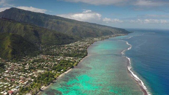 French Polynesia, Moorea island vibrant coastline with dense populated villages and residential areas, green mountains and an expansive turquoise lagoon with visible coral reefs. Aerial drone flight
