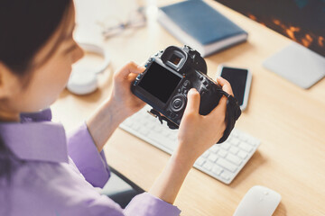 Naklejka premium Focused Young Woman Holding Digital Camera And Inspecting Photos At Home Office, Seated At Desk Surrounded By Creative Tools And Modern Gadgets. Engaging In Photography Concept