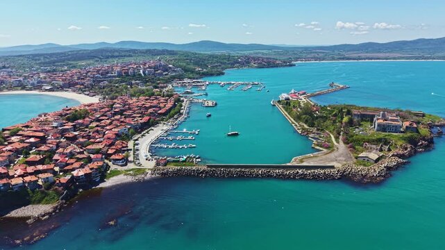 Nessebar shows the harbor filled with boats and the beach area along the coast. The town is surrounded by hills under a clear blue sky.